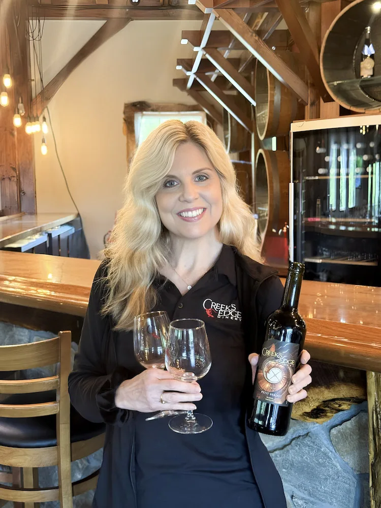 A woman holding a bottle of wine and two wine glasses at Creek's Edge Winery.